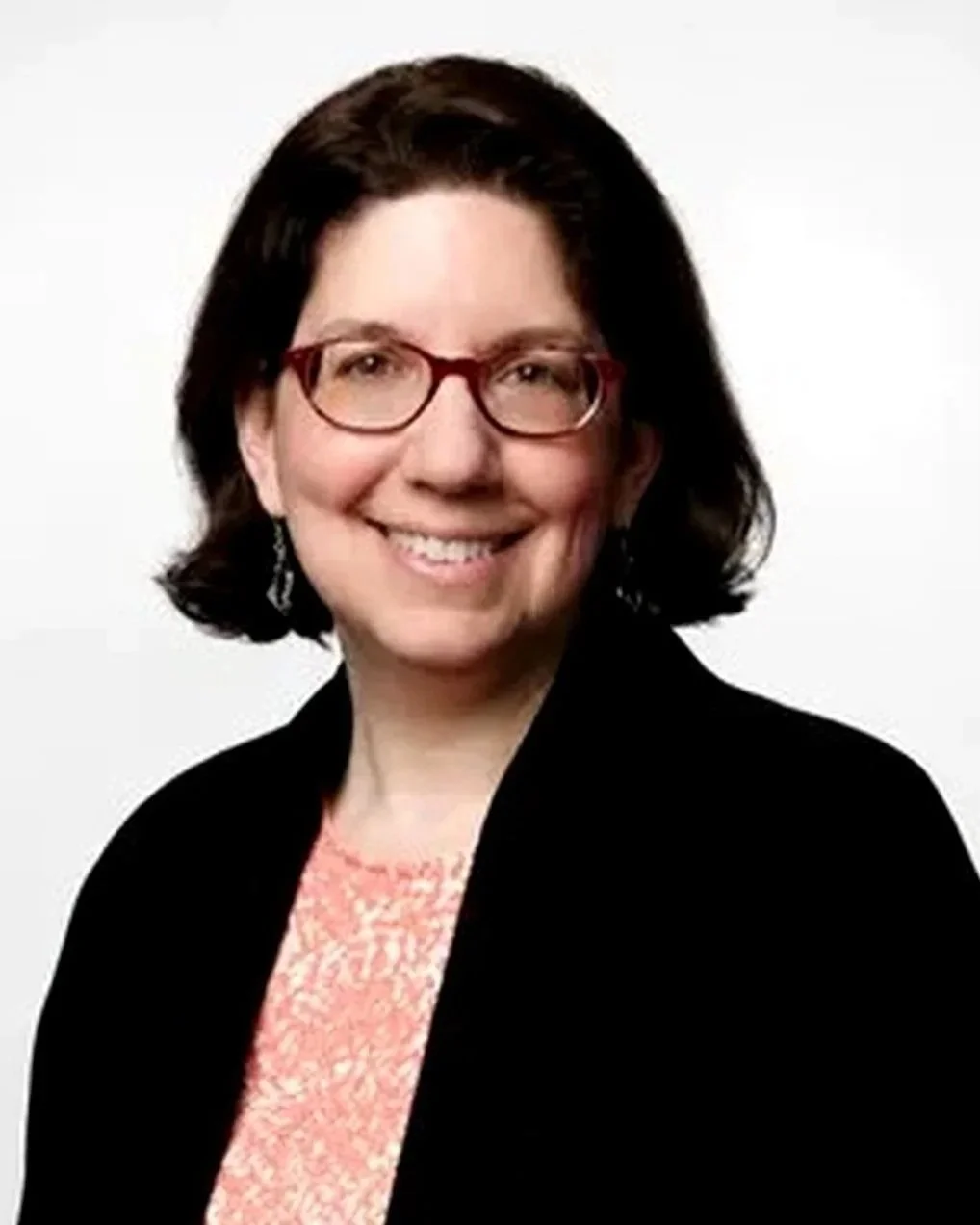 A woman with short dark hair, wearing glasses, a black blazer, and a pink patterned blouse, smiling against a white background.
