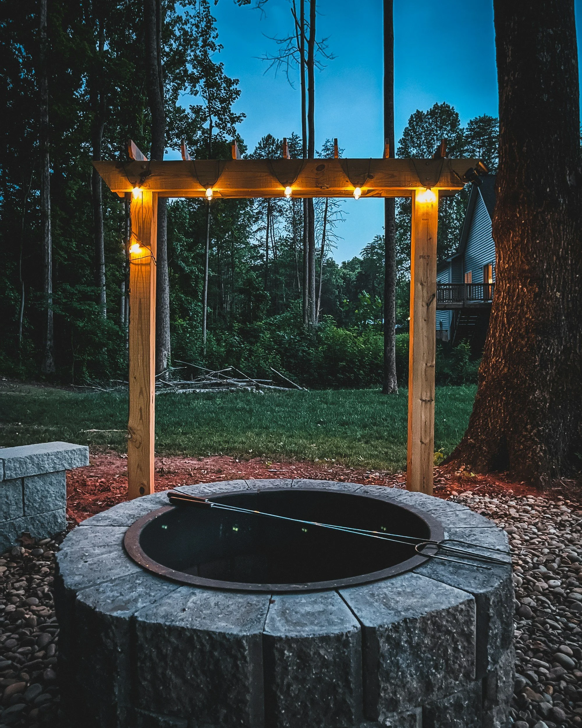 A fire pit made of stone with a metal ring inside, situated outdoors on a gravel surface, with a wooden arbor with string lights hanging from it, surrounded by trees at dusk.