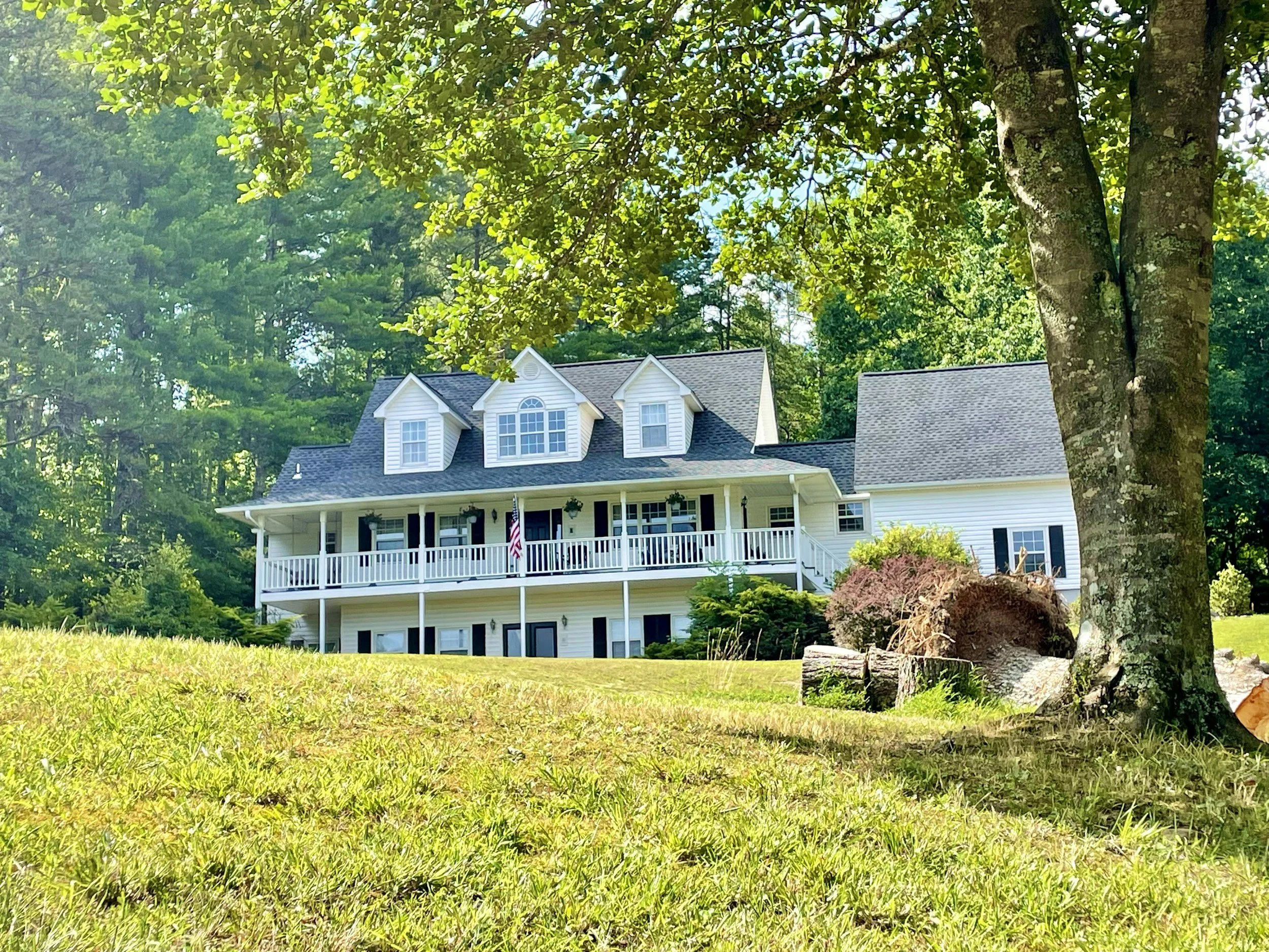 A white house with a porch and dormer windows, surrounded by green trees and a lawn, under a bright sky.