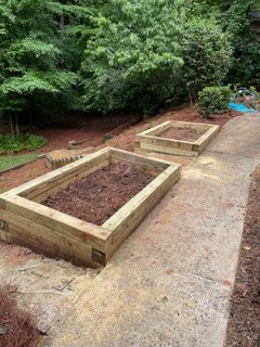 Three newly built wooden garden beds filled with soil on a paved surface, with trees and bushes in the background.