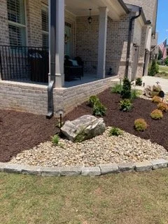 A front yard landscaping scene with a small garden bed bordered with gray bricks, filled with mulch, rocks, small bushes, and a large decorative rock, beside a brick house with a porch.
