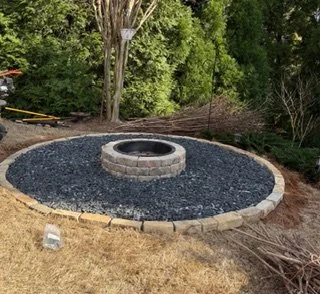 A circular fire pit area with brick edging, filled with black gravel, surrounded by grass and trees.