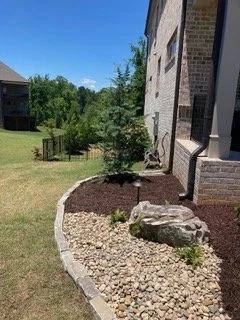 A landscaped yard with a curving stone border, small plants, and a mulch bed next to a house with brick and siding exterior.