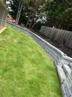 A sloped backyard with a green lawn and a curved gray retaining wall made of concrete blocks, surrounded by trees and a wooden fence.