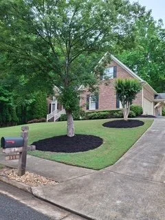 A brick house with a sloped roof, surrounded by a well-manicured lawn with three newly planted trees with mulch around their bases, and a driveway leading to the garage.
