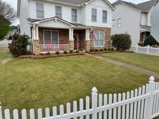 Front yard with a grassy lawn, white picket fence, and a house with a porch and bushes.