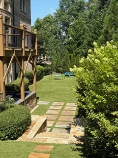 Backyard with a wooden deck, stone pathway, green lawn, bushes, and trees, with a house to the left.