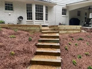Wooden stairs leading up to a house with a white brick exterior and a garden area with sparse plants.