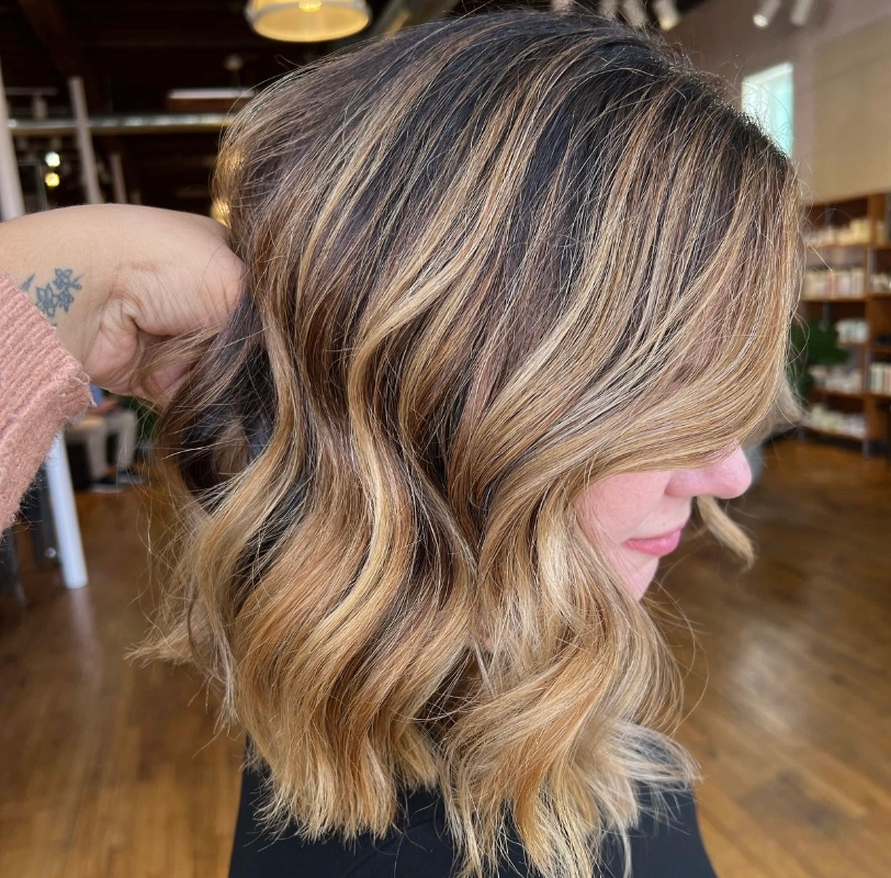 Close-up of a woman with shoulder-length, wavy, blonde hair with darker roots, inside a salon or studio with wooden flooring and shelves in the background.