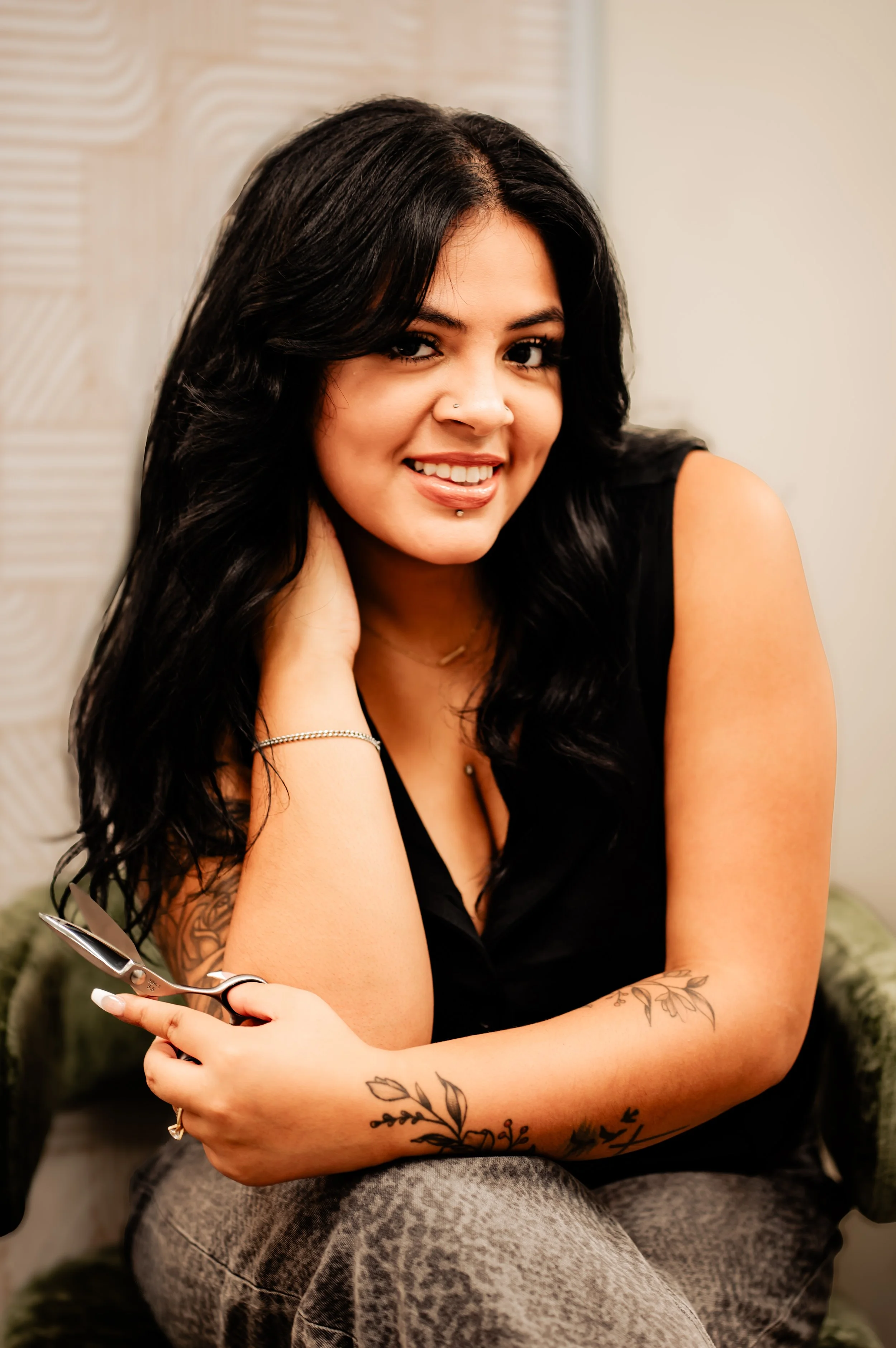 A woman with black hair, tattoos on her arms, holding scissors, sitting and smiling in a casual setting.