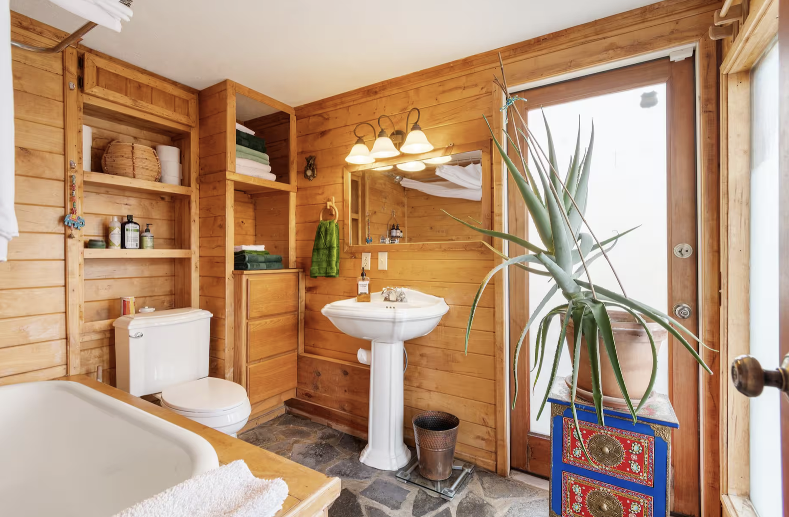 Bathroom with wood-paneled walls, a pedestal sink, and a large plant in a pot near a door with frosted glass. Shelves with towels and toiletries are visible, and a mirror above the sink reflects part of the room.
