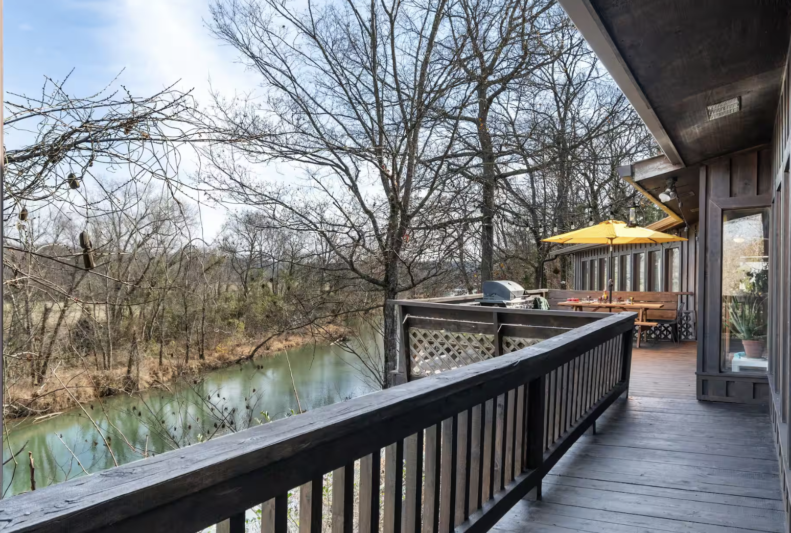 A wooden balcony overlooking a river, with leafless trees in the background and a yellow patio umbrella on the deck.
