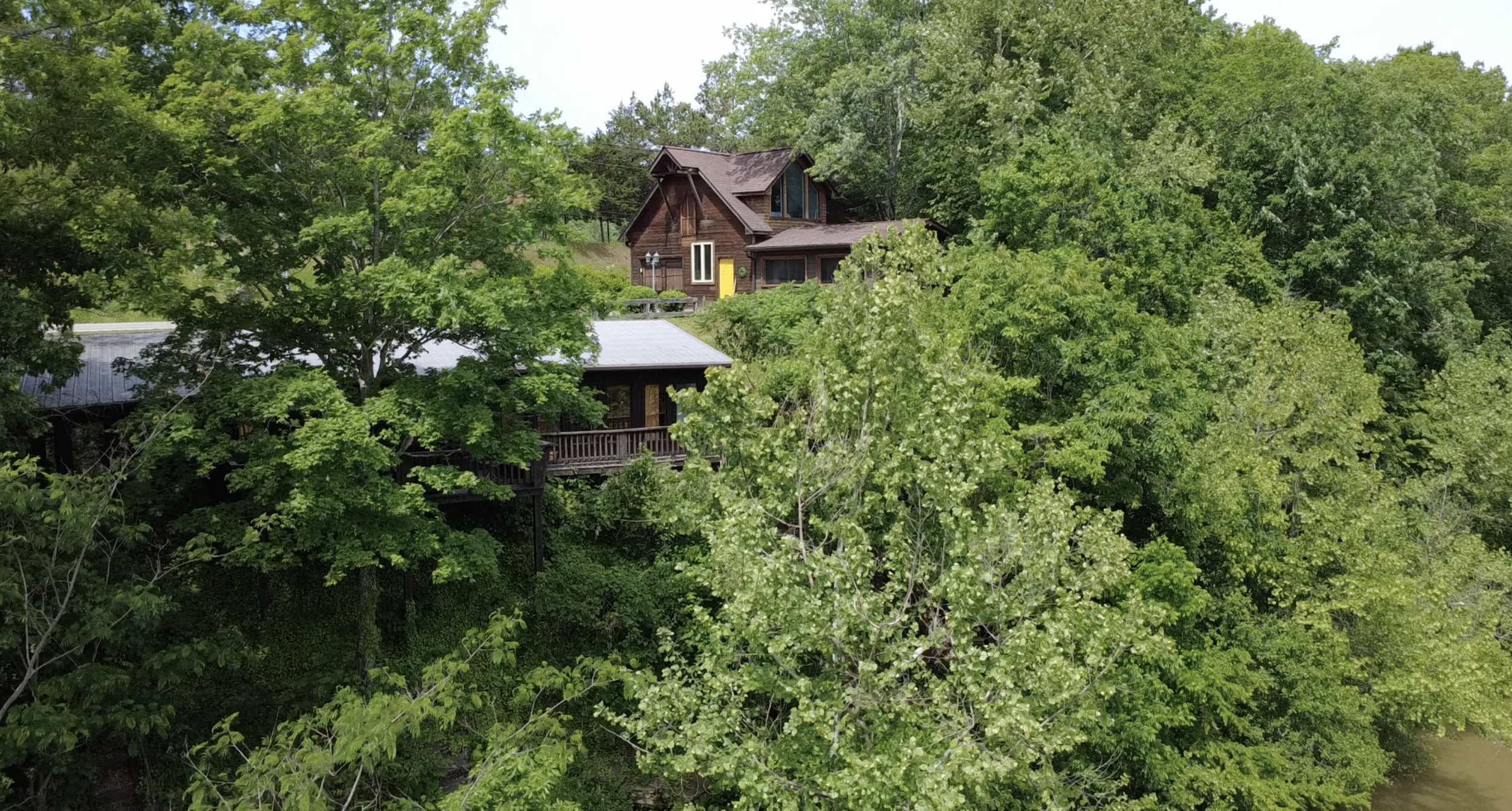 A wooden house with a steep roof and large windows is perched among dense green trees near a waterway.