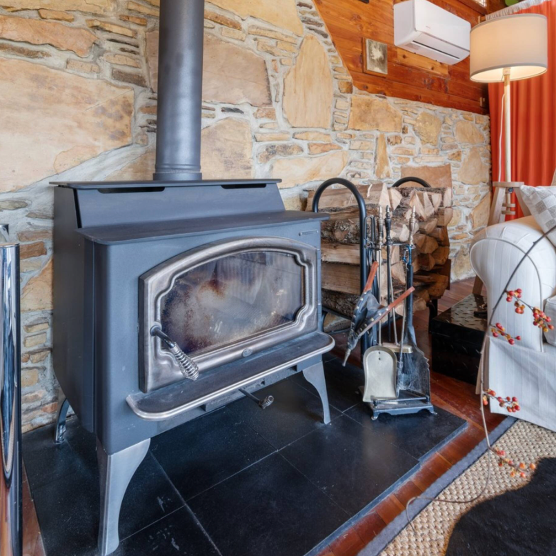 A cozy living room corner featuring a wood-burning stove on a black tiled hearth, with a stone wall behind and fireplace tools next to it. To the right, there is a stack of firewood, a white armchair with a floral pillow, a floor lamp, and a window with orange curtains.
