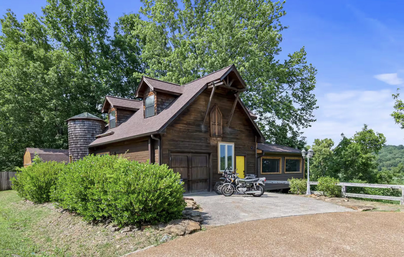A rustic barn-style house with dark wooden exterior, yellow door, and small dormer windows, surrounded by green trees and bushes, with a driveway and parked motorcycle and scooter.
