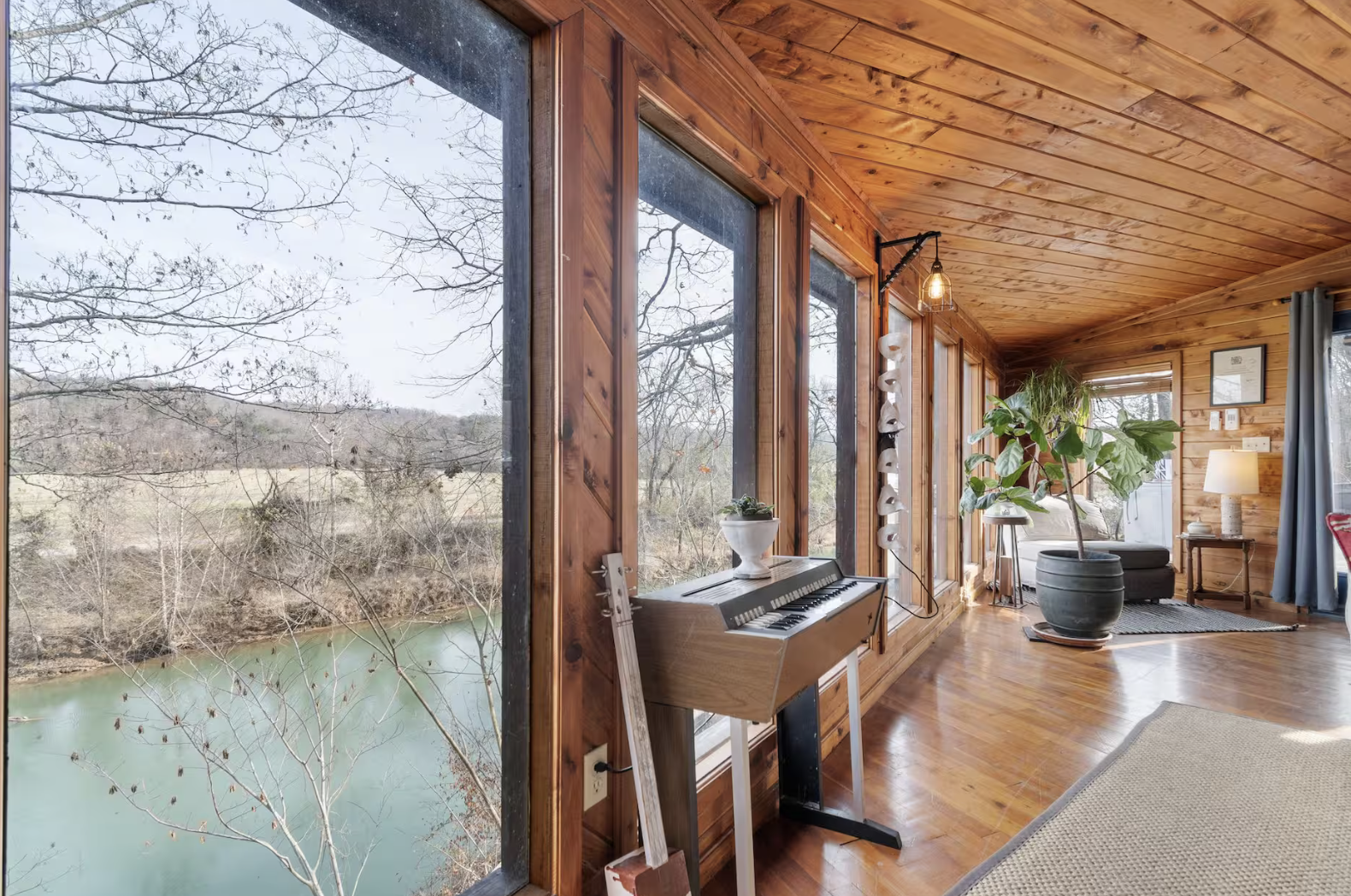 A cozy, sunlit room with large windows offering a view of a river and bare trees outside. The interior features wooden walls and ceiling, a digital keyboard on a stand, a potted plant, a small side table with a lamp, and a comfortable sitting area with an armchair and curtains.