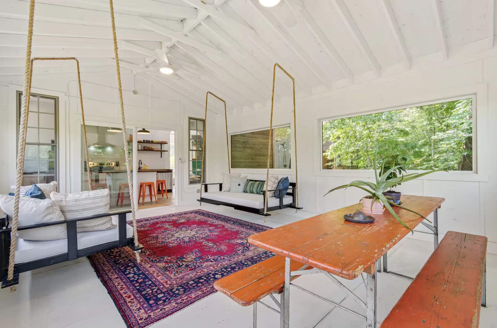 Bright living room with white walls and ceiling, featuring a red patterned area rug, white swings with ropes, a wooden table with a potted plant, and large windows showing green trees outside.