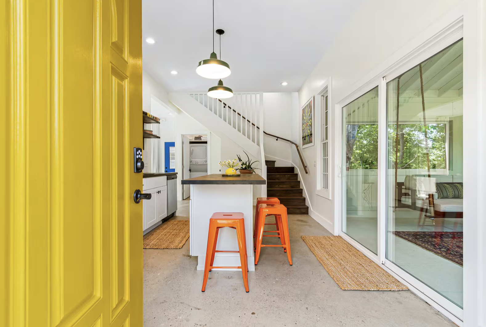 Interior view of a modern house with a yellow front door, kitchen area, stairs, and a living room visible through large glass sliding doors, featuring orange bar stools, plants, and artwork.
