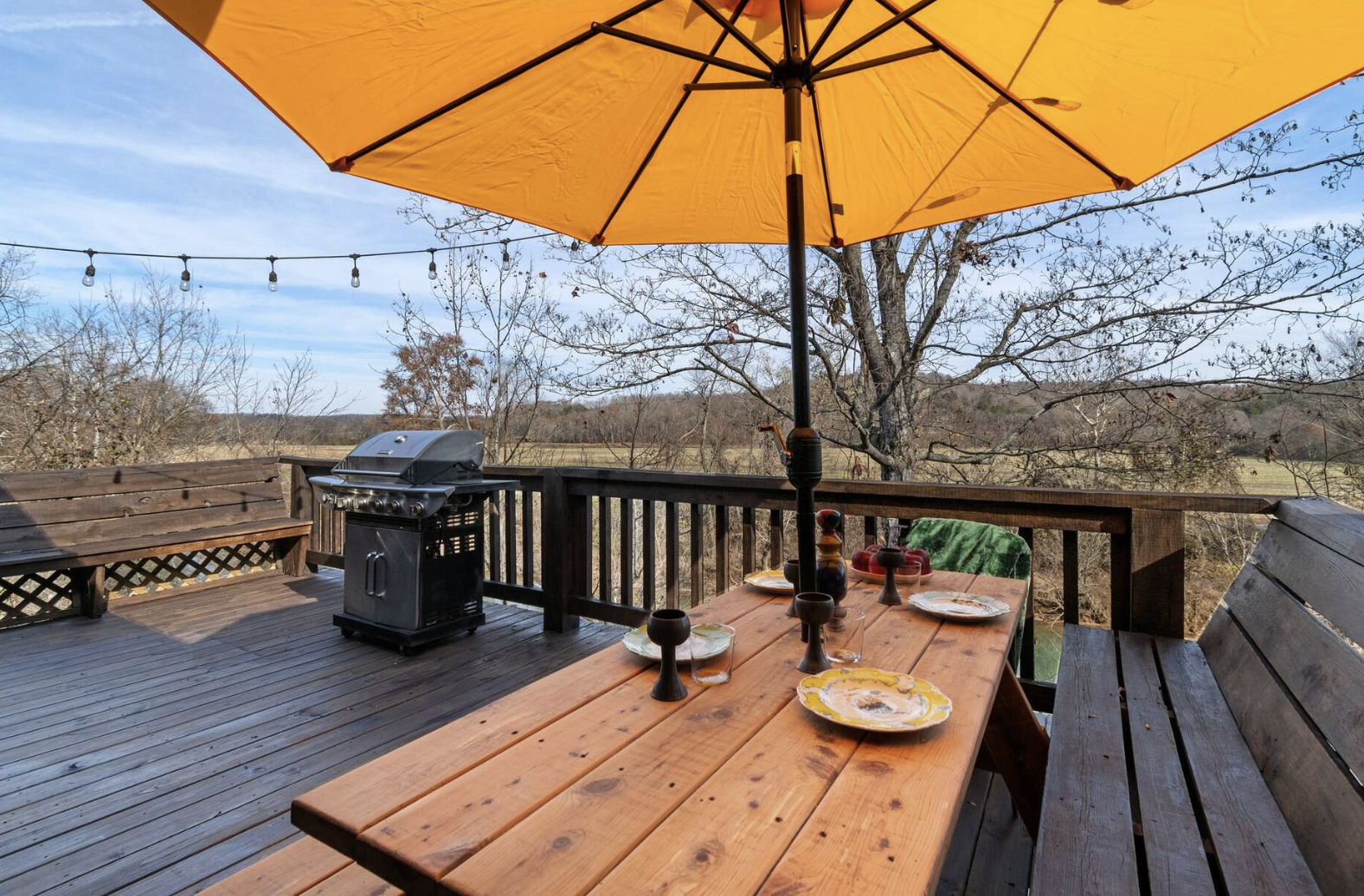 Outdoor wooden deck with a table set for dining, a large yellow umbrella, a barbecue grill, and string lights, overlooking a scenic landscape with trees and open fields under a blue sky.