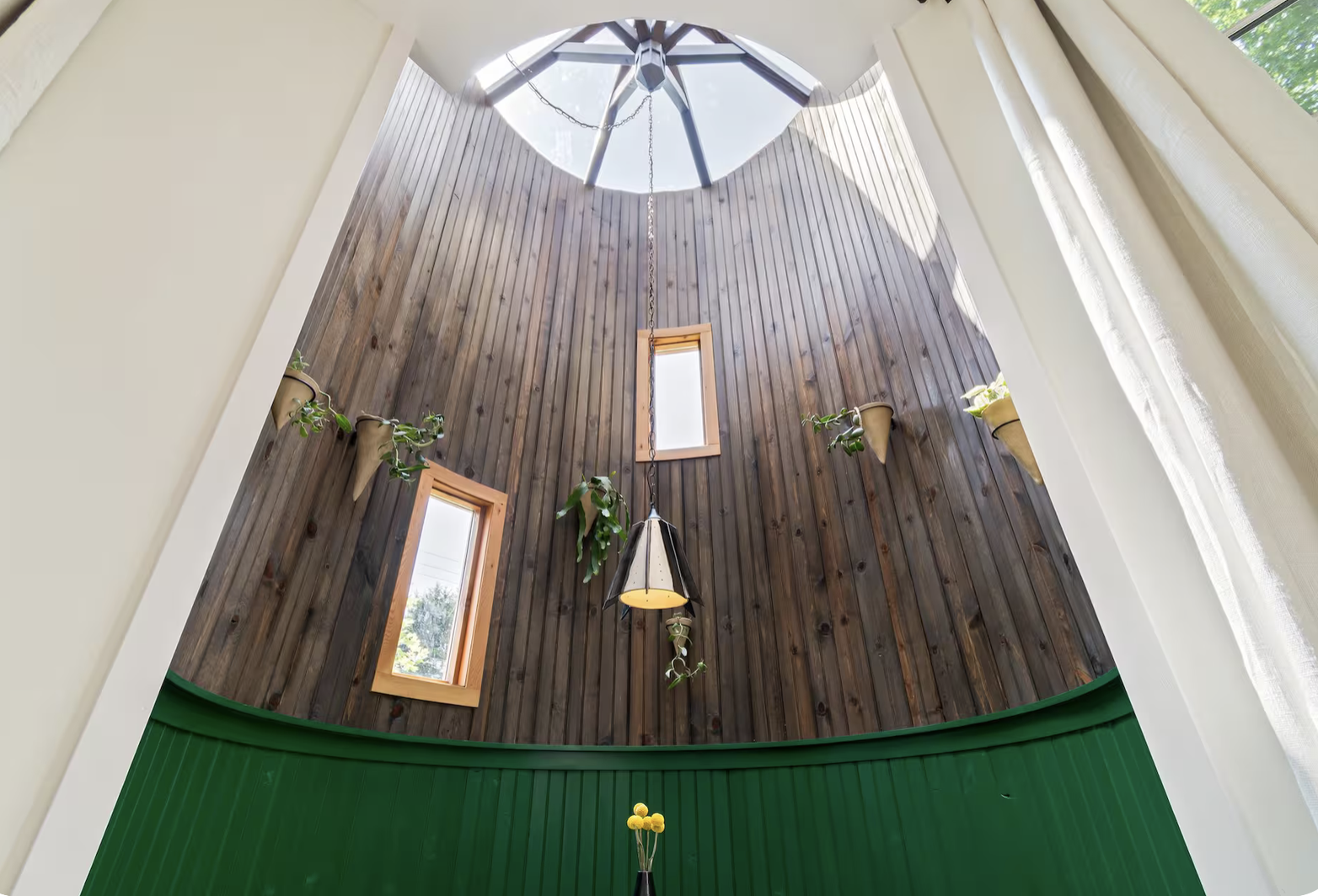 Interior view of a high ceiling with a wooden wall, two small windows, potted plants, a hanging light fixture, and a glass skylight at the top, with curtains on the sides.