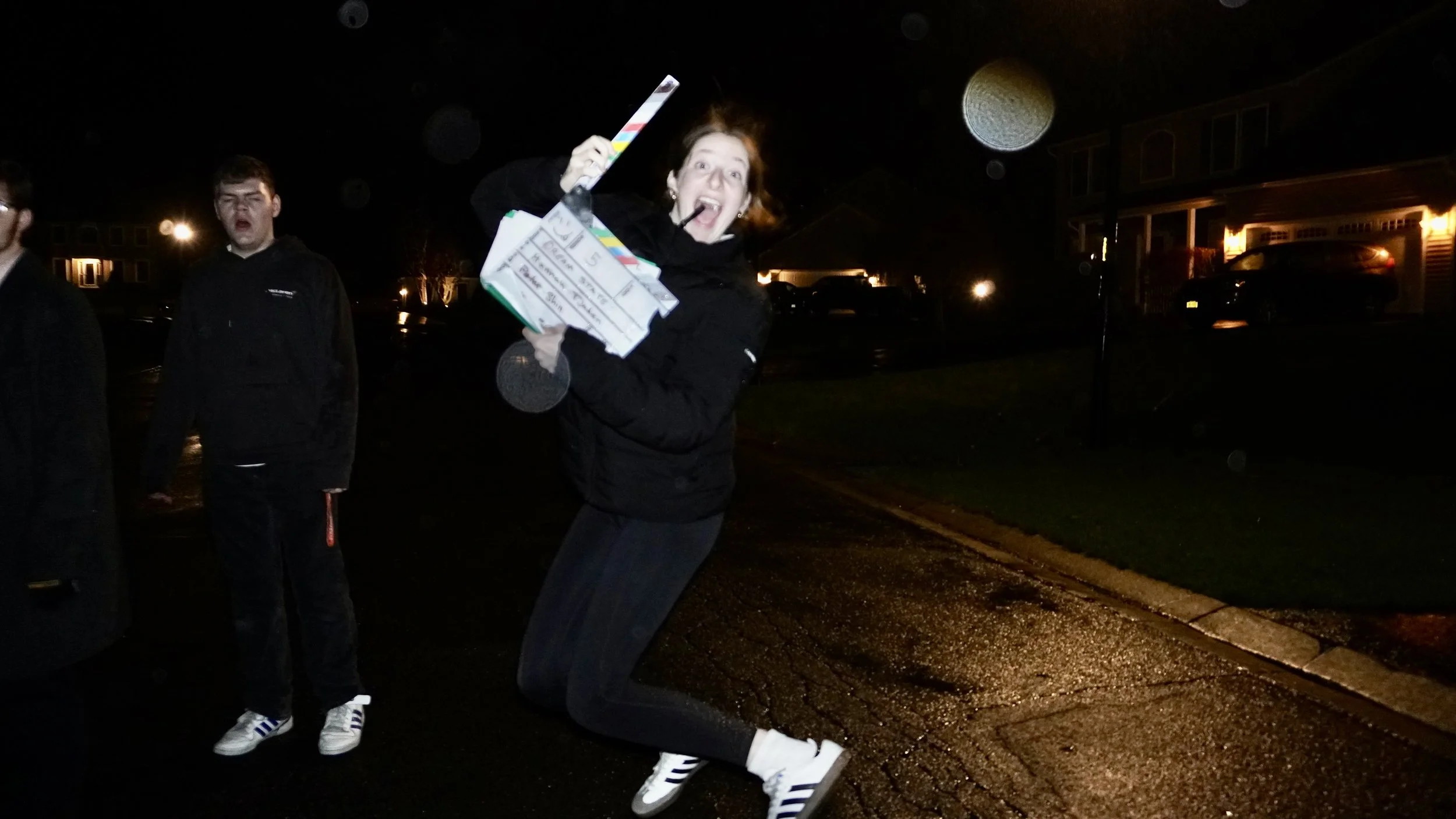 A woman joyfully holding a clapperboard in a parking lot at night, with her mouth open in excitement and two young men standing nearby, all dressed in dark clothing.