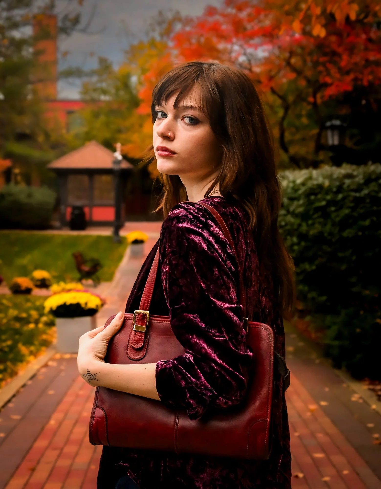 A young woman with brown hair and blue eyes standing outdoors in autumn, holding a maroon handbag, with colorful fall trees and a brick pathway in the background.