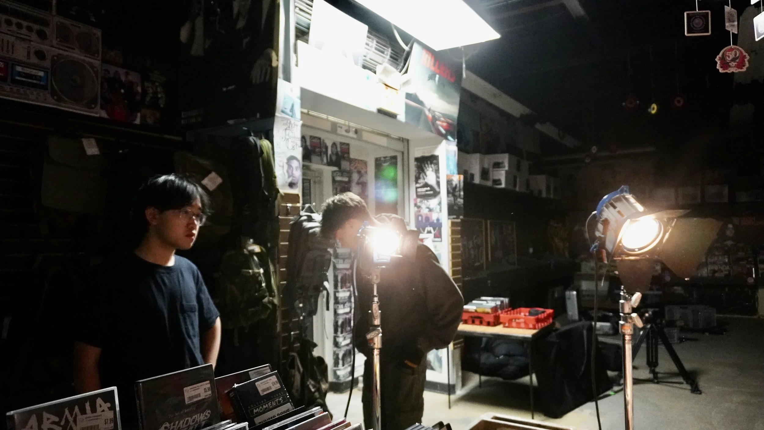 People working on a film or photography set inside a store or shop, with lighting equipment and magazines displayed in the foreground.