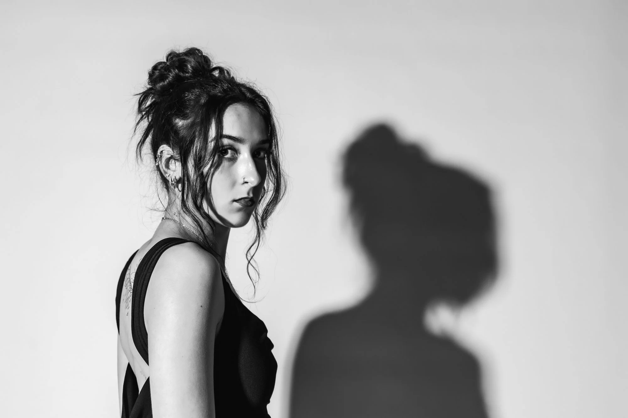 A young woman with curly hair tied in a bun, wearing a sleeveless black top, looking over her shoulder in a black-and-white photo with a shadow on the wall behind her.