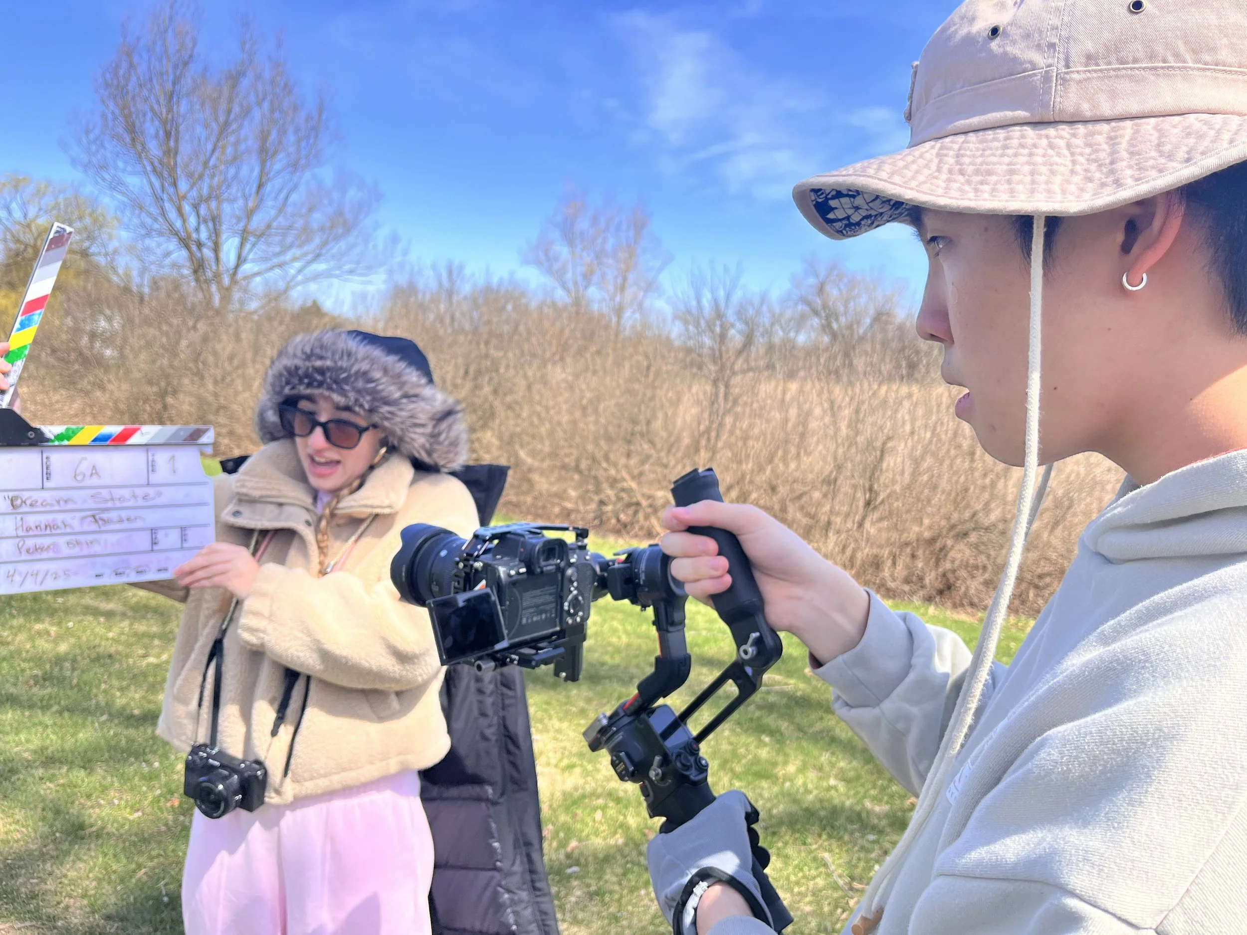 Two young women outdoors, one holding a clapperboard with text, and both with cameras and equipment, under a blue sky with trees in the background.