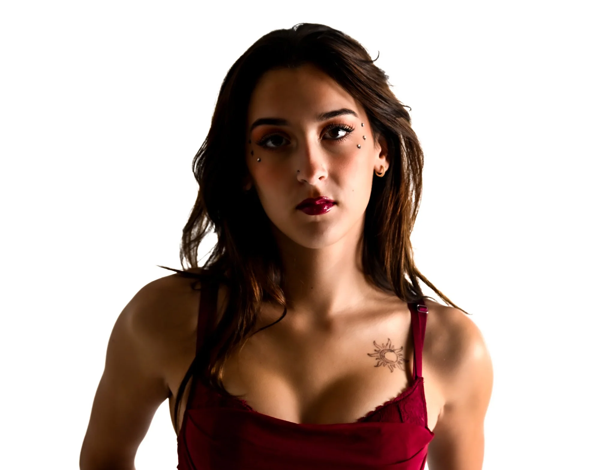 A young woman with medium-length brown hair and multiple facial piercings, wearing a burgundy lace camisole top, standing against a white background, with a sun tattoo on her chest.