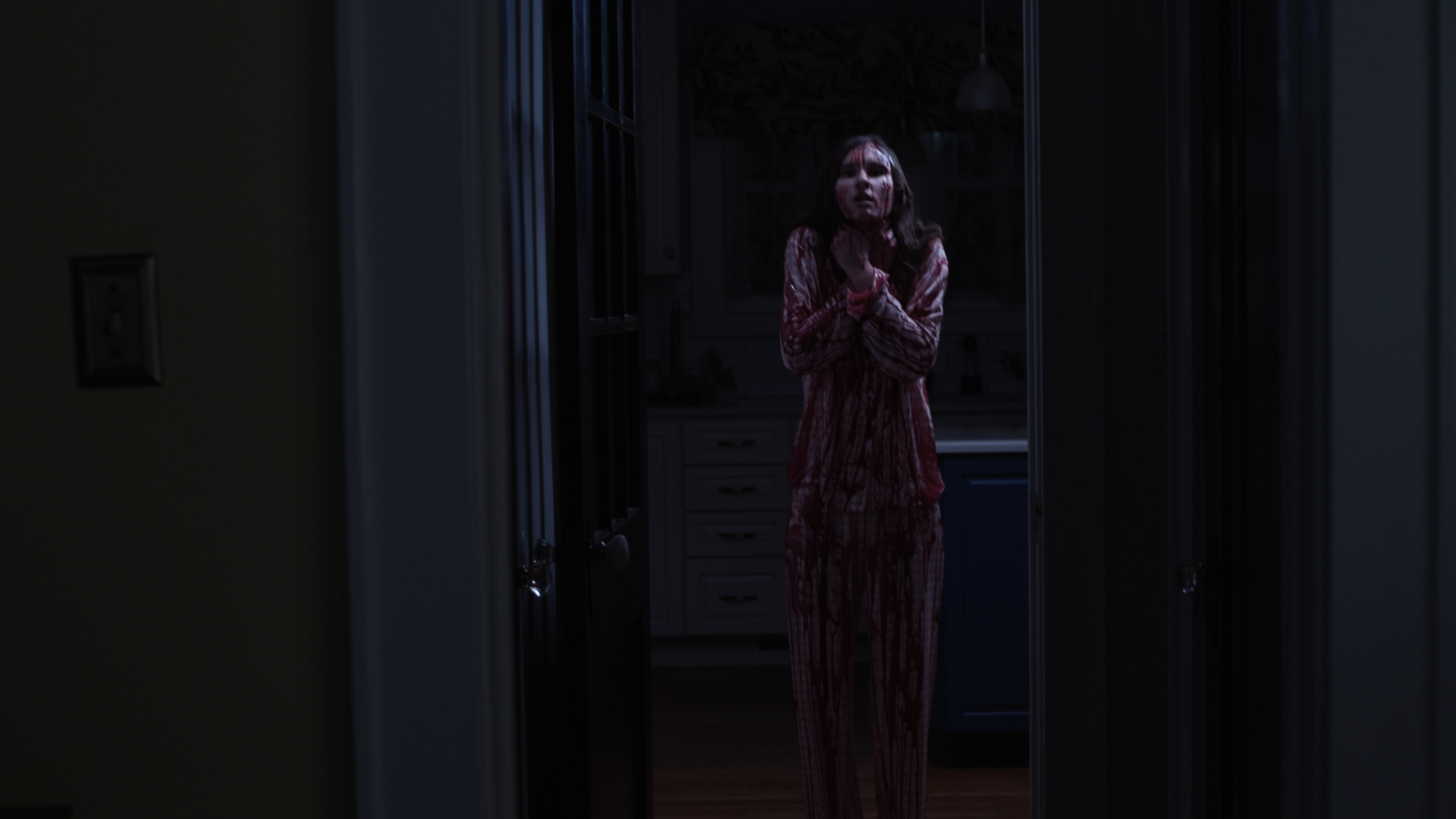 A woman standing in a dark kitchen, covered in blood, wearing pink pajamas, looking frightened or shocked.