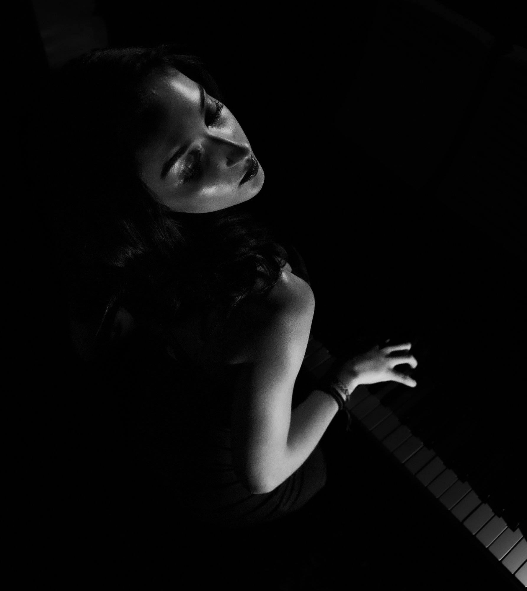Black and white photo of a woman with closed eyes, long wavy hair, and makeup, playing a keyboard in a dark setting.