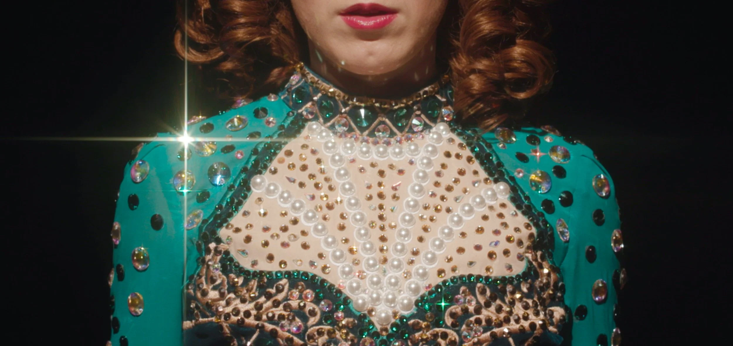 Close-up of a person wearing an elaborate, jewel-studded costume with sequins, pearls, and gemstones, with only the lower half of their face visible against a black background.