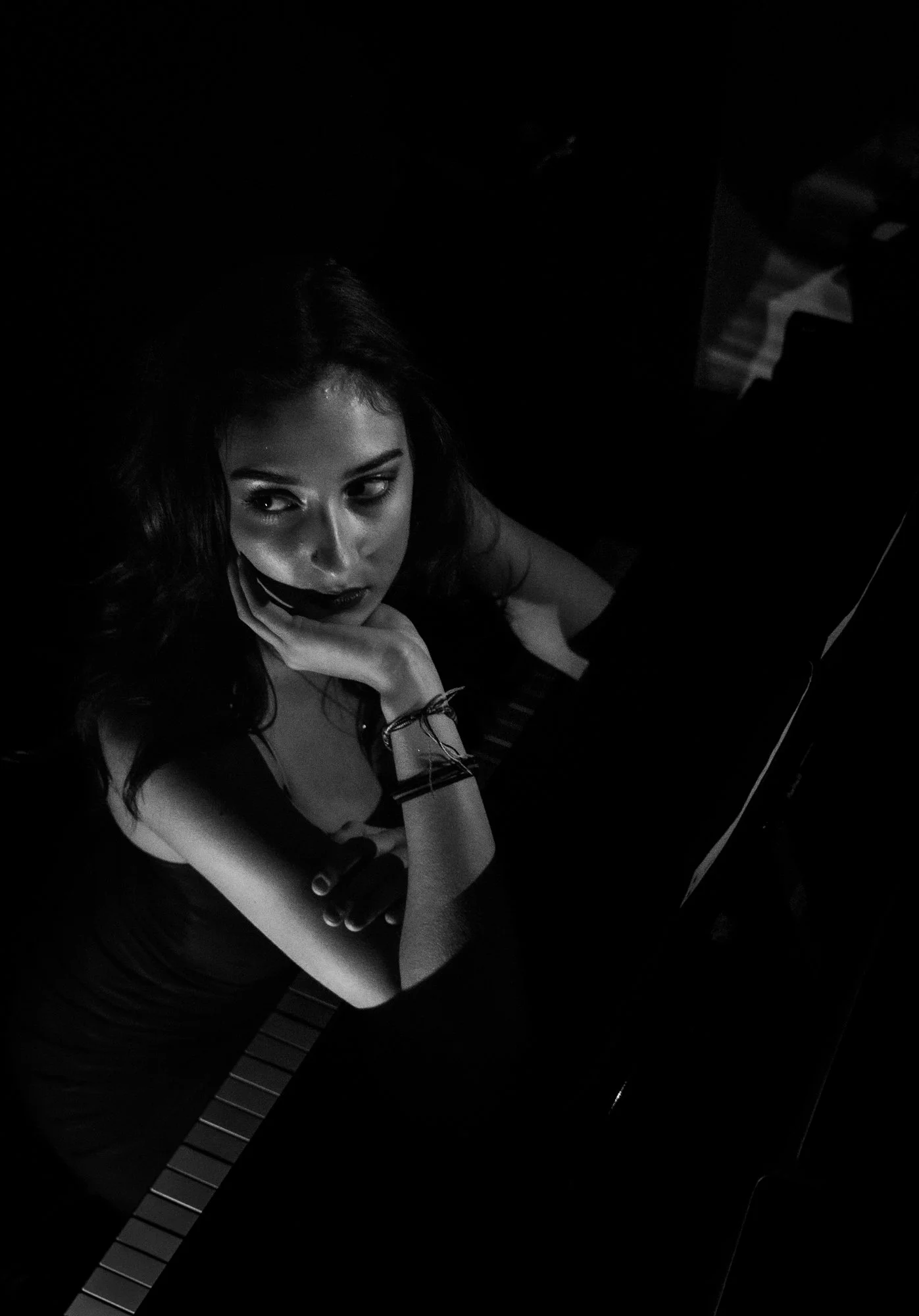 A woman with dark hair, wearing dark lipstick, sitting at a piano with her hand resting on her chin, looking to the side, with a dark background.