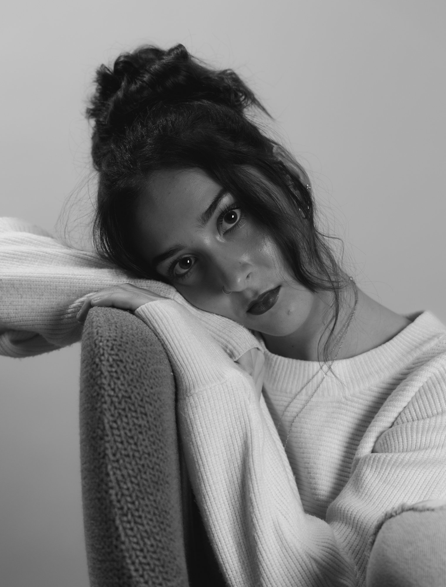 A black and white photo of a woman resting her head on her arm, looking directly at the camera. She has dark, wavy hair and is wearing a white, ribbed long-sleeve shirt, with a neutral-colored blanket or fabric draped over her arm.