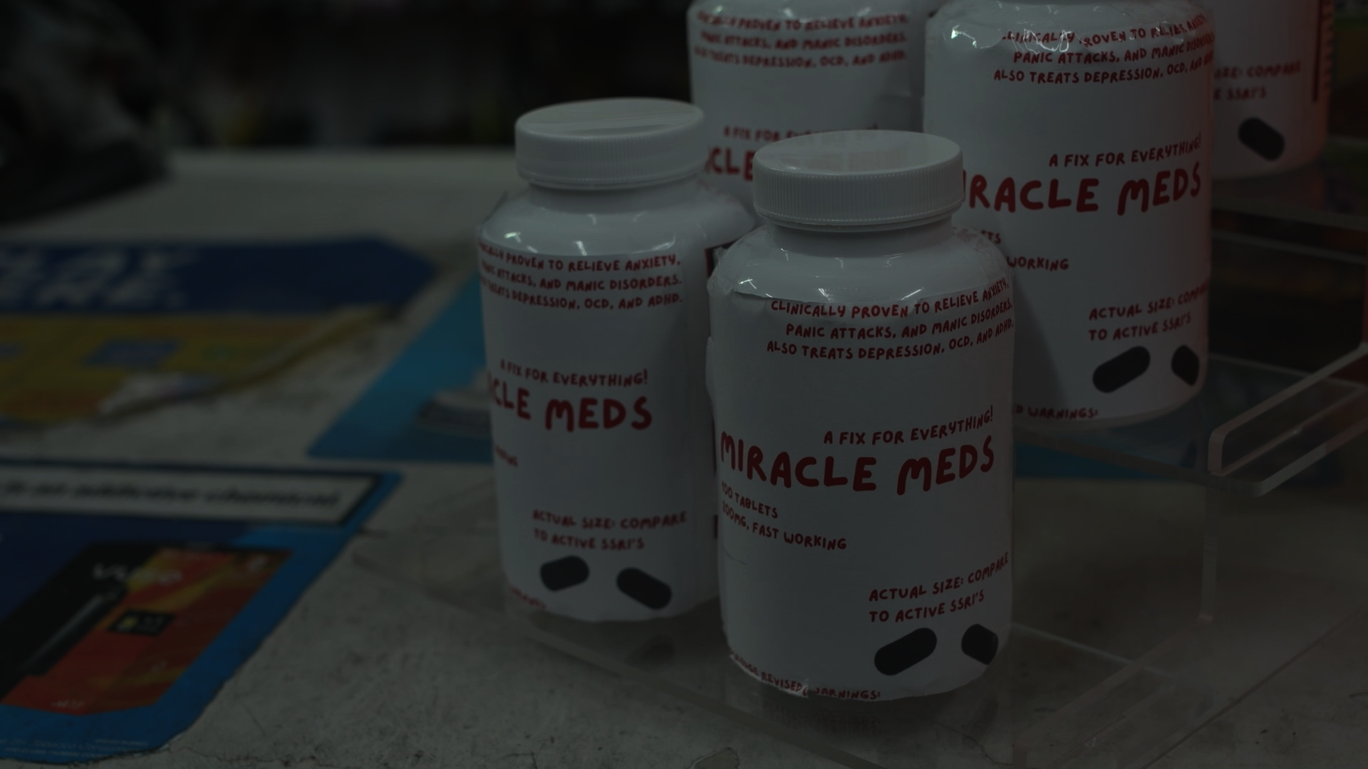 Bottles of Miracle Meds with white labels and red text, surrounded by other pharmacy items and brochures on a counter.