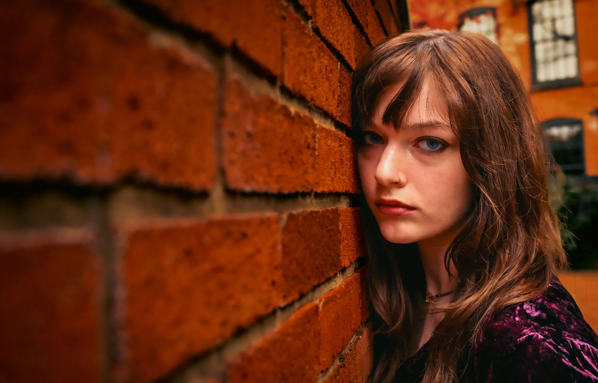 A young woman with brown hair and blue eyes peeking out from behind a brick wall, wearing a dark floral top.