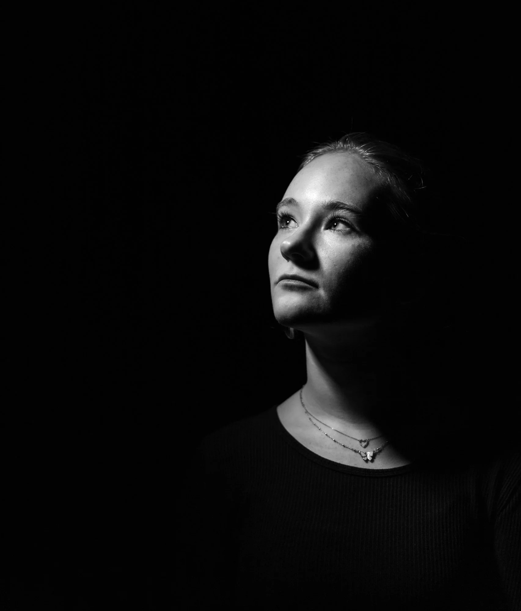 Black and white portrait of a young woman looking upwards, wearing layered necklaces, with her hair pulled back, against a dark background.