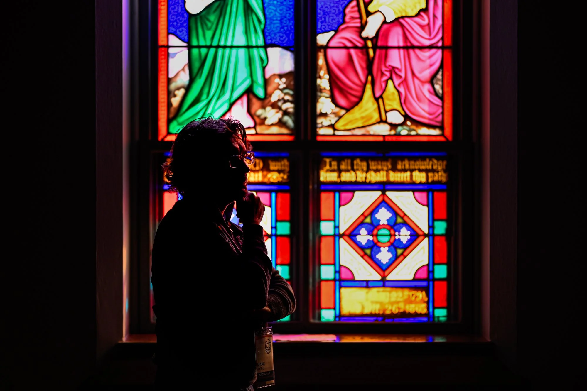 Person with glasses standing in front of a stained glass window with colorful biblical artwork, including figures dressed in robes.