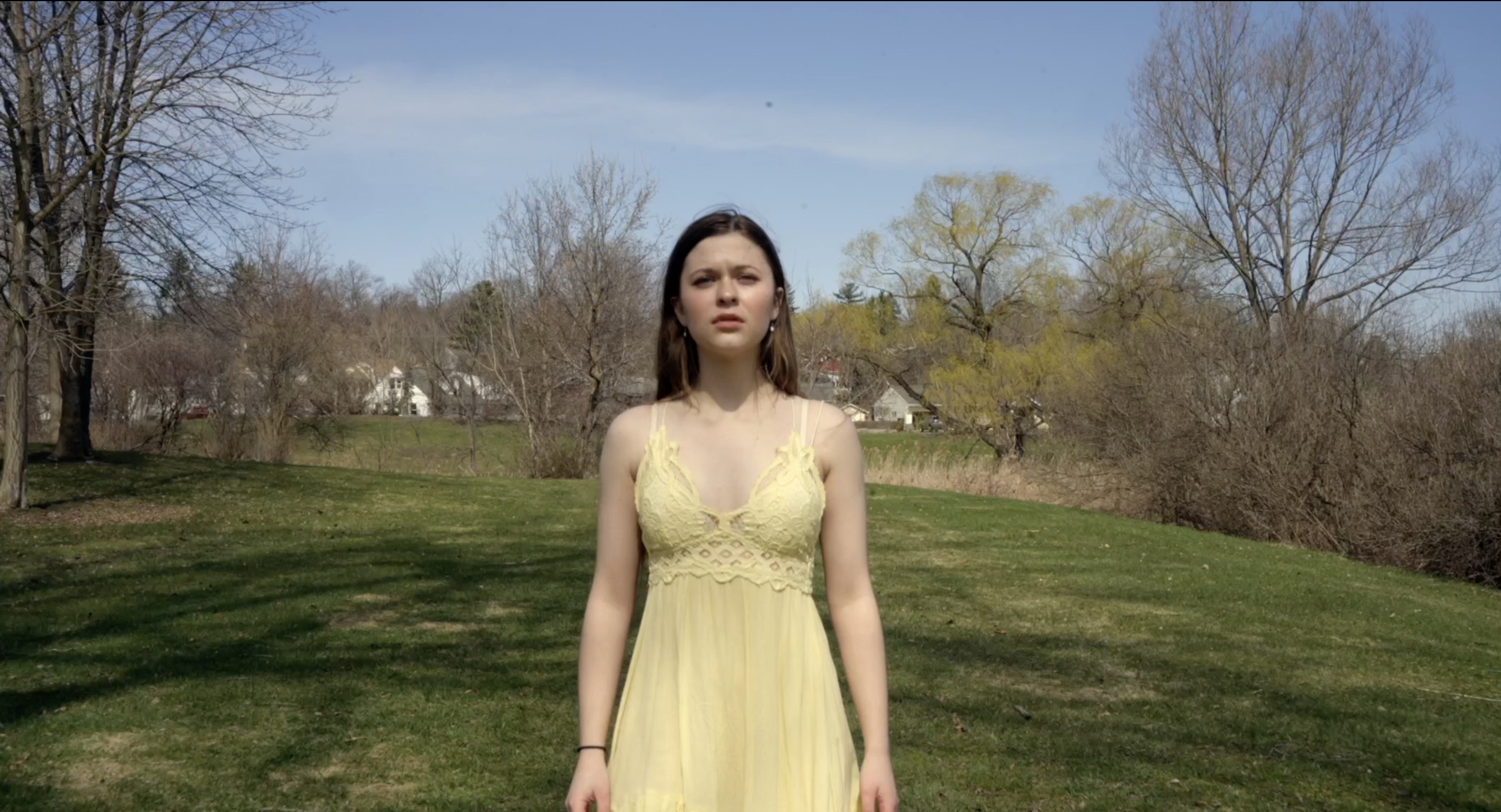 A young woman stands in a park with trees and houses in the background, wearing a yellow dress and looking at the camera.