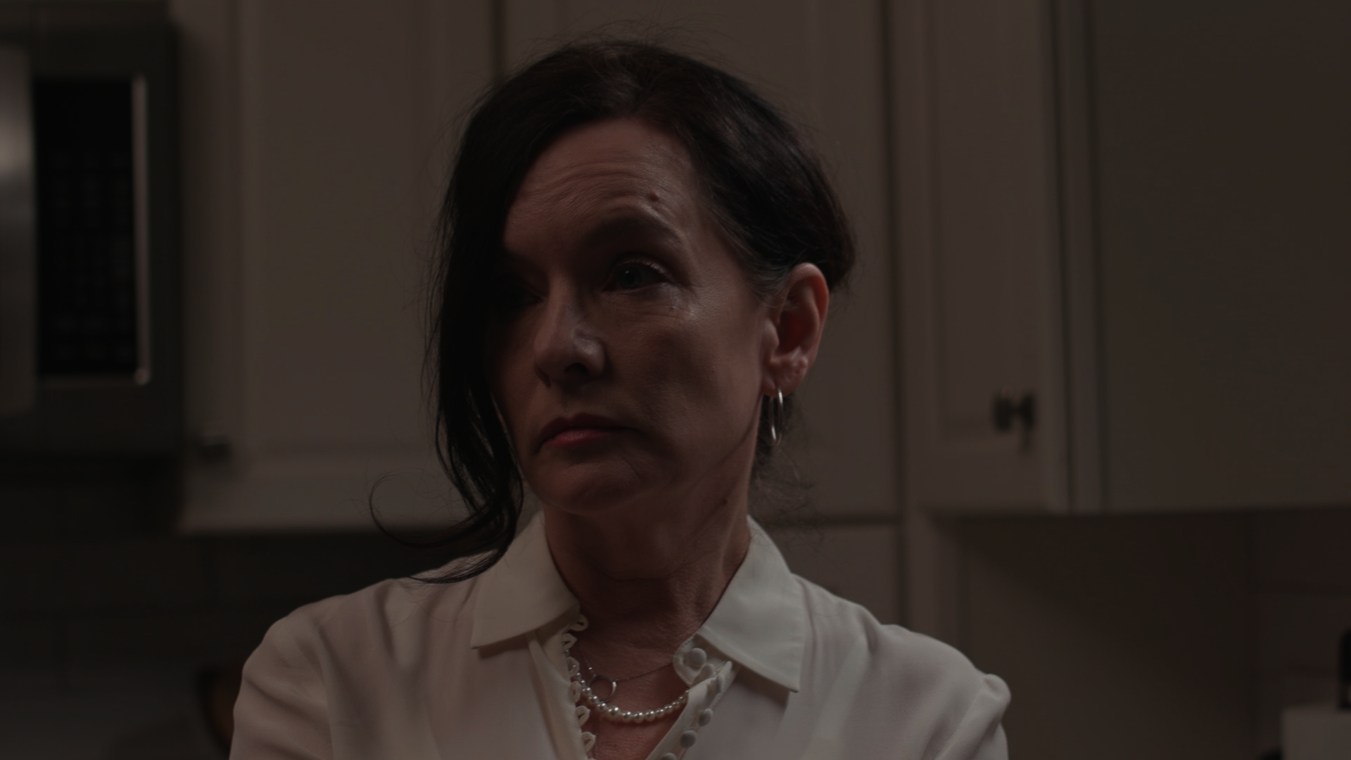 A middle-aged woman with short, dark hair, wearing a white blouse and pearl jewelry, stands in a kitchen with beige cabinets and a microwave in the background, looking to her right with a serious expression.