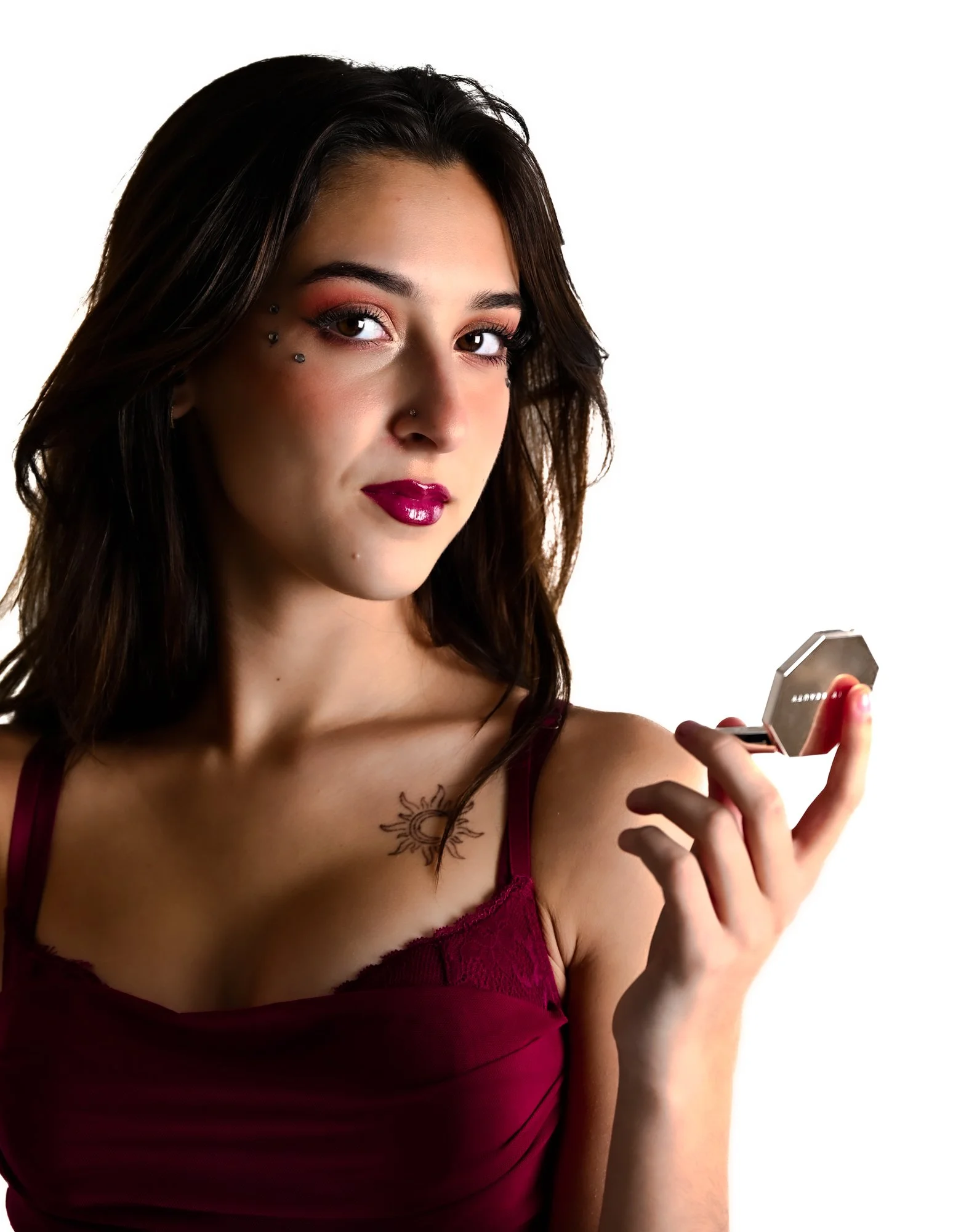 A young woman with dark hair and makeup, wearing a burgundy camisole, holding a compact mirror, and looking at her reflection.