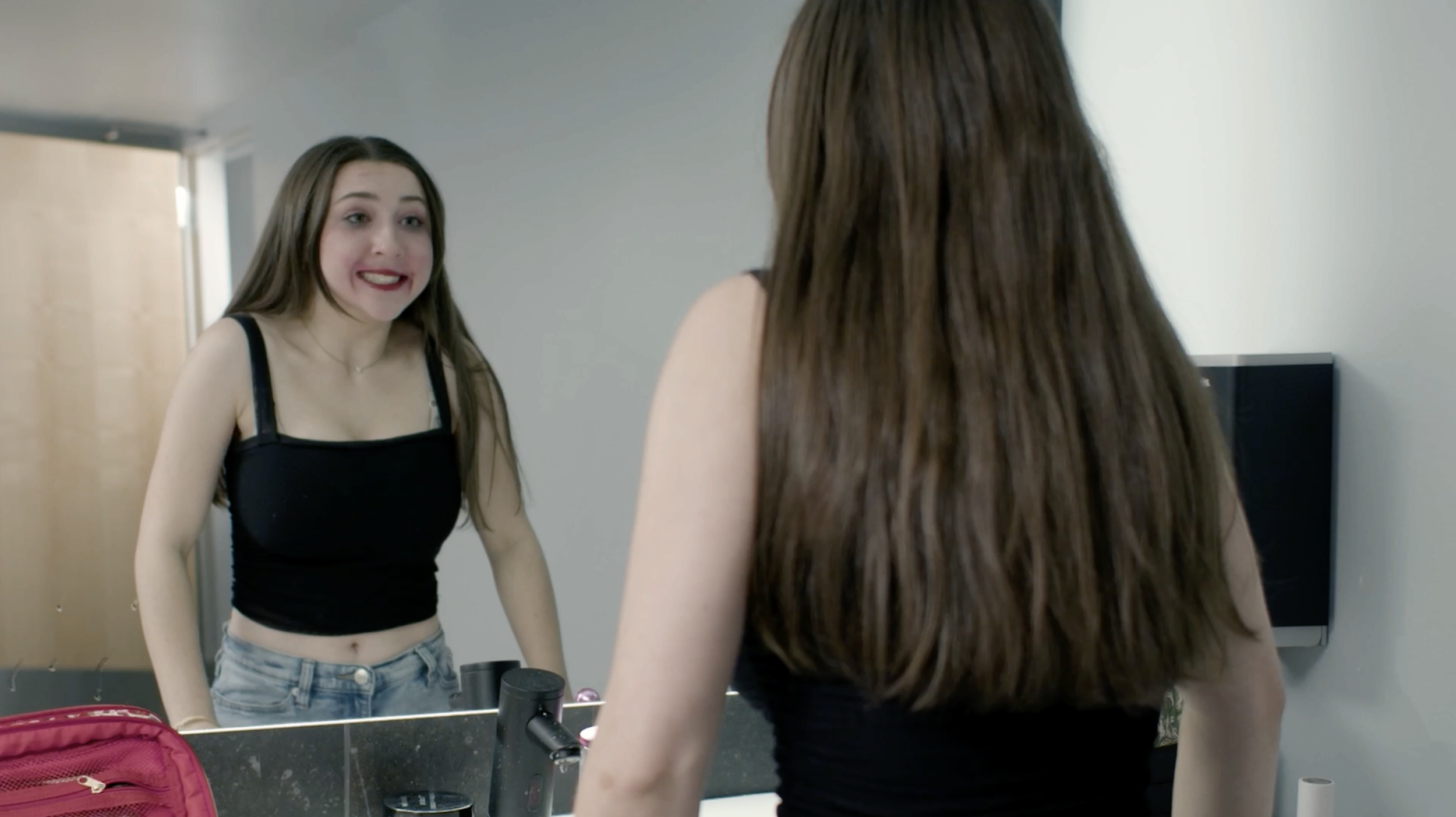 A young woman with long brown hair and a black tank top looking at herself in a mirror in a bathroom or dressing room, with a red bag, makeup items, and a sink visible.