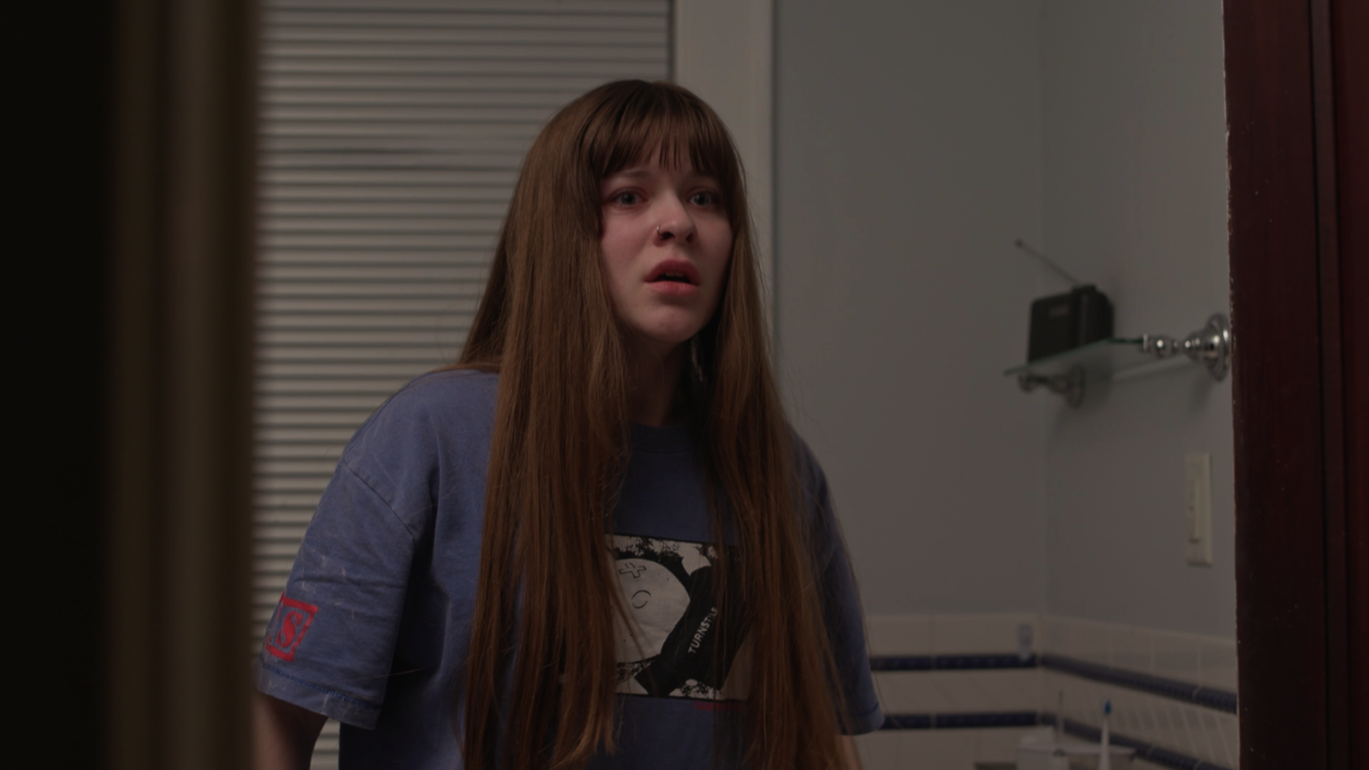 A young woman with long brown hair and a nose piercing looks surprised in a bathroom with a white wall, wooden door, and a shelf with a radio.