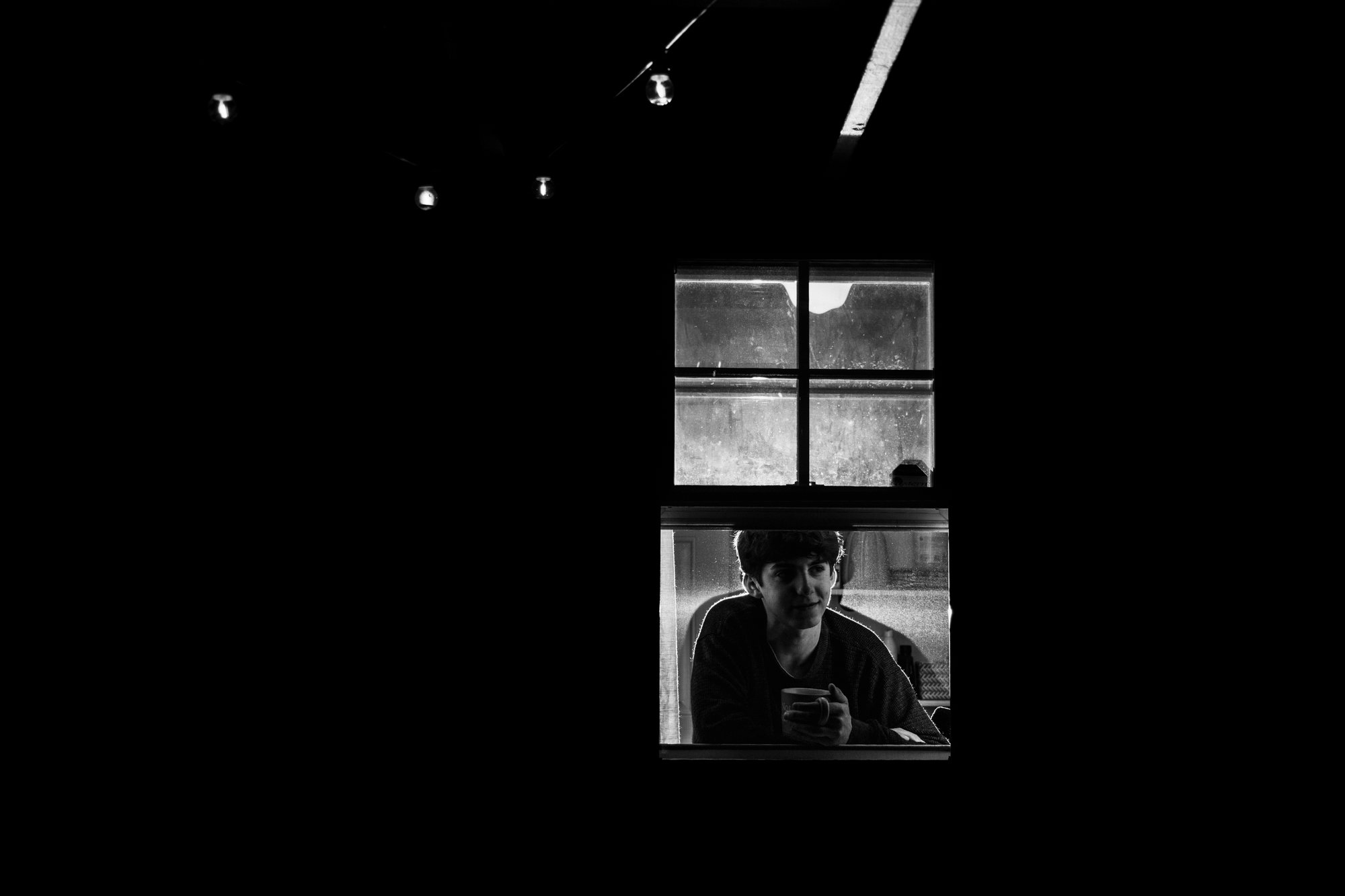 A young man with dark hair looking out of a window, holding a mug in his hand, with a dimly lit room behind him and a cloudy sky outside.