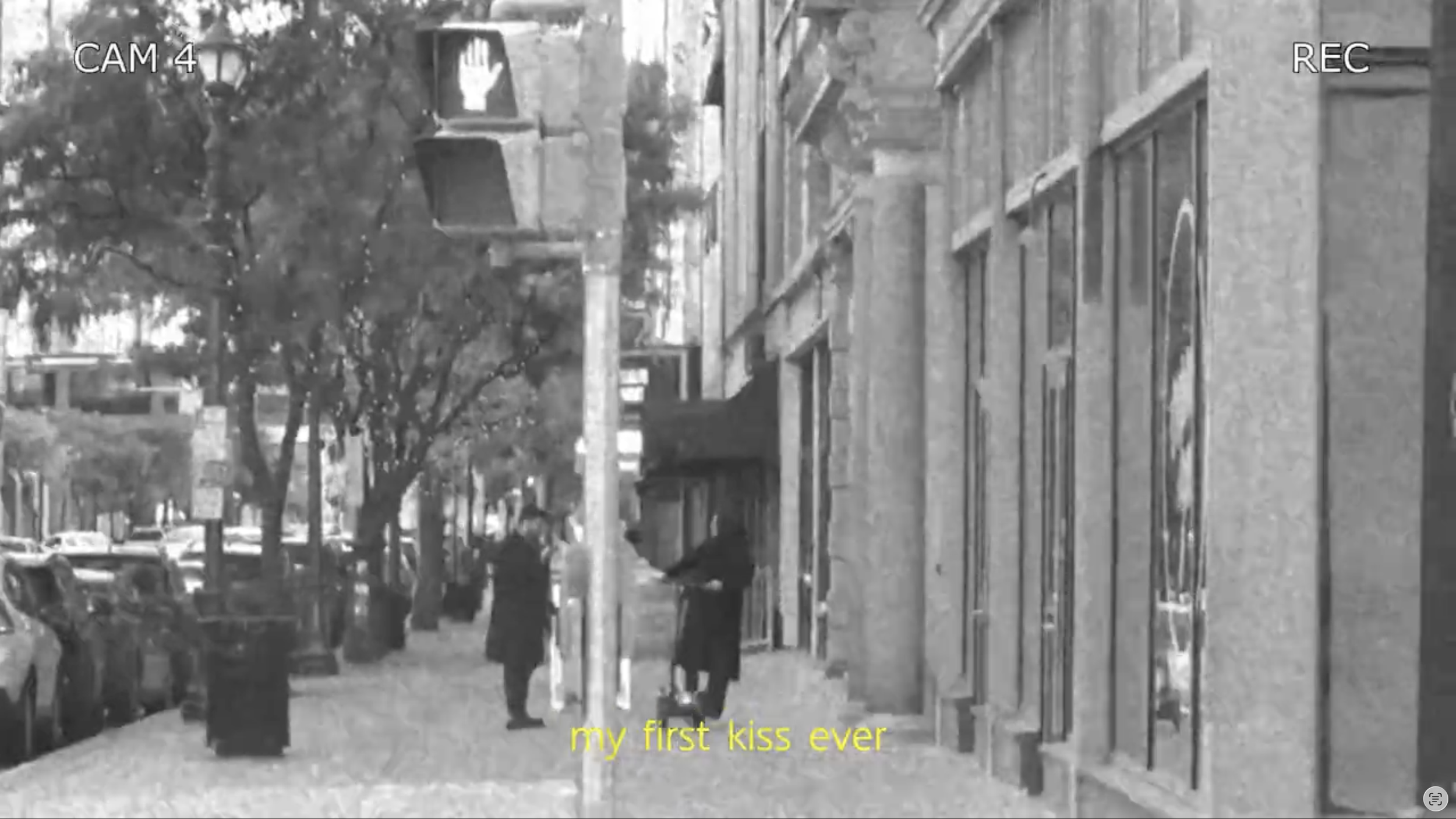 A street scene in black and white with a couple walking together under trees, in front of shops and parked cars. There are street lamps, trees, a utility pole, and a building with windows. The caption at the bottom reads 'my first kiss ever.'