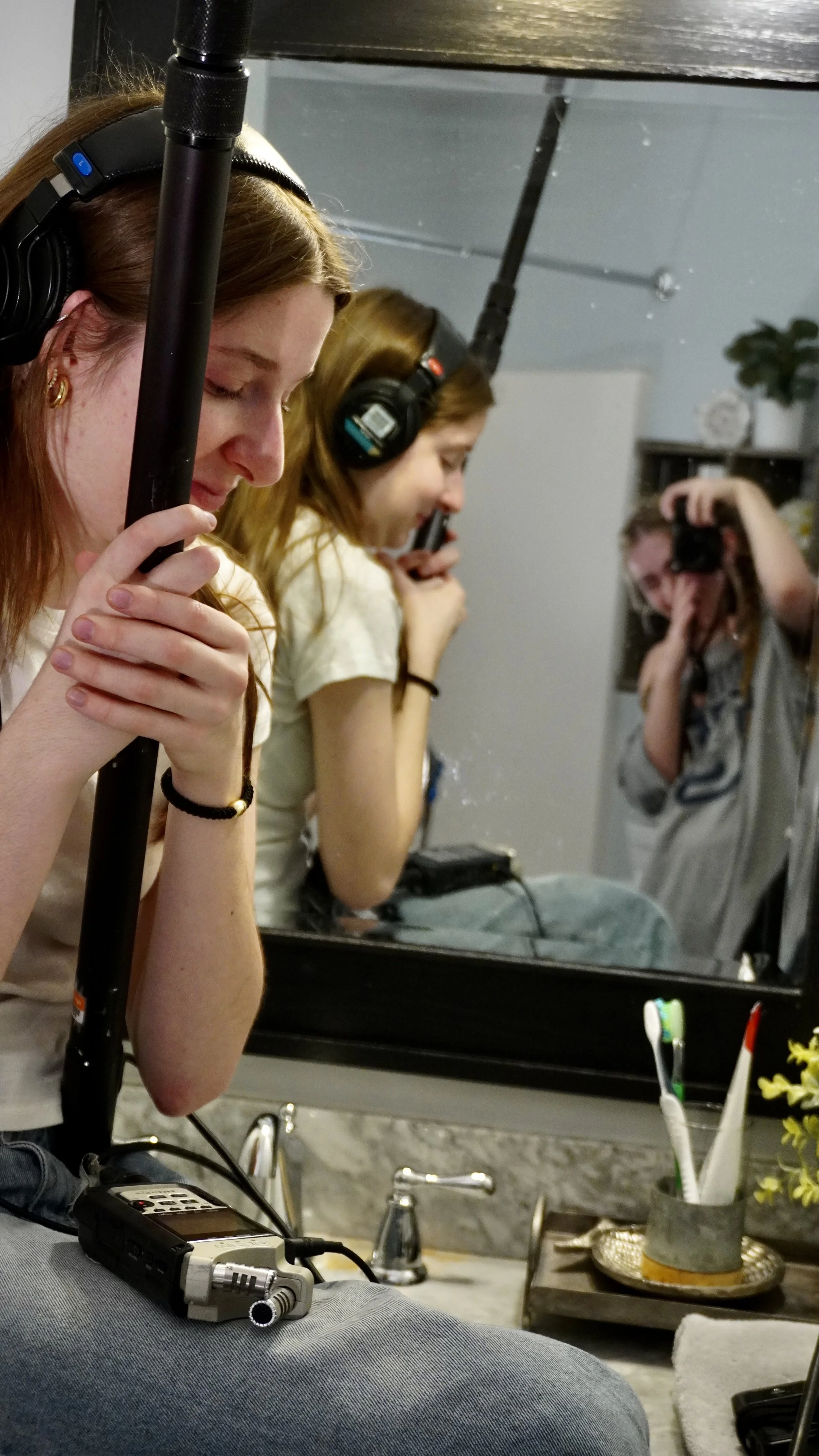 Three women with red hair and wearing headphones sit by a mirror with their eyes closed, seemingly meditating or resting. The background includes a sink, a counter, and a shelf with decorative items, including a plant.