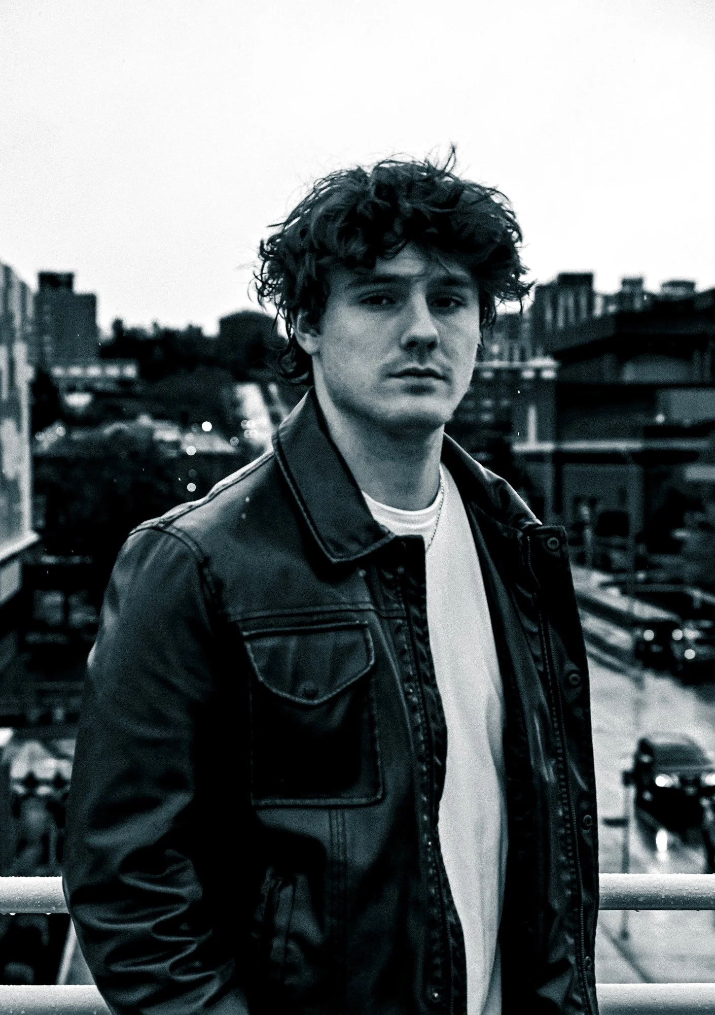 A young man with curly dark hair wearing a leather jacket and white t-shirt, standing outdoors against an urban rooftop background.
