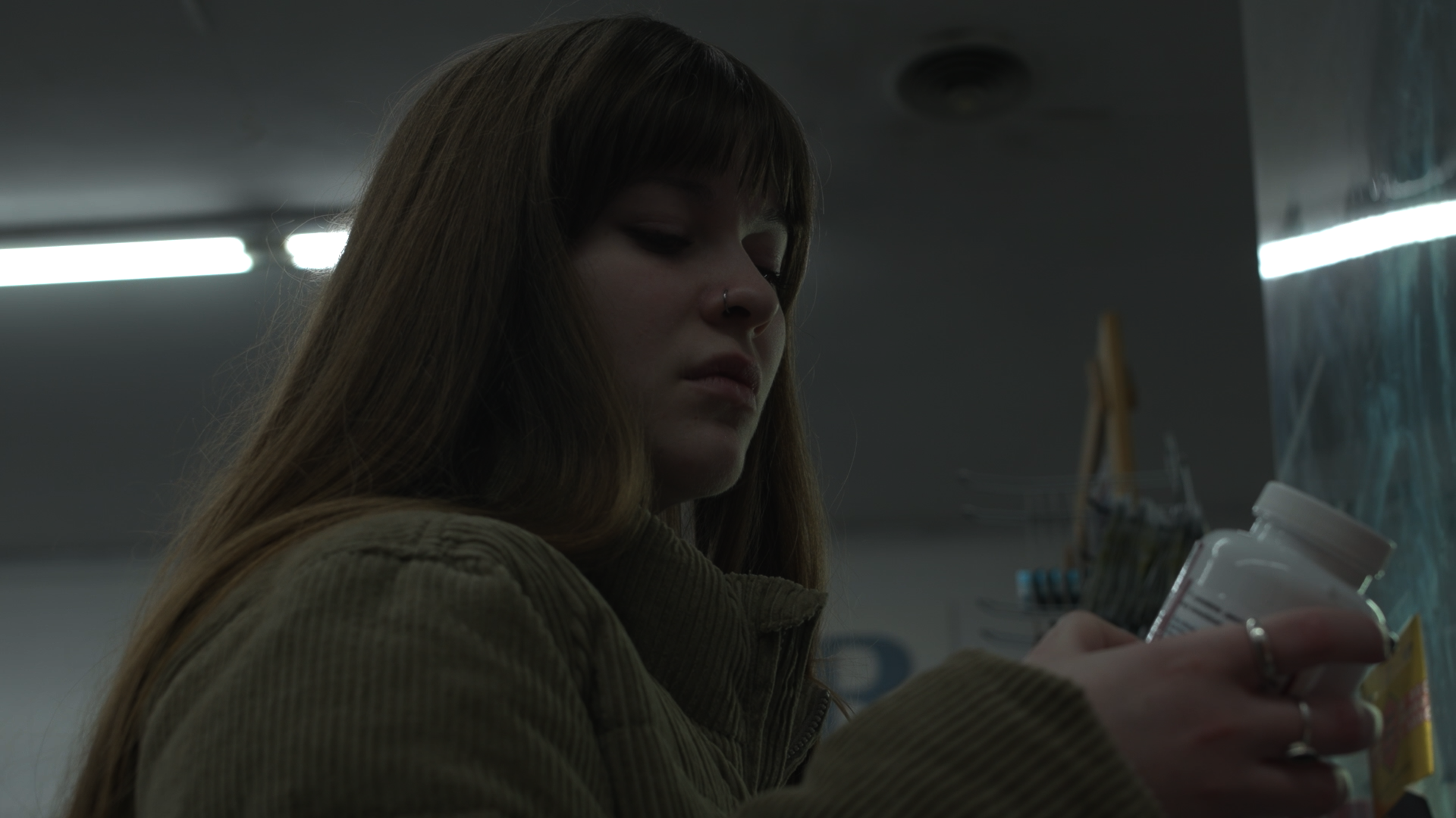 A young woman with long brown hair and a nose piercing looks at prescription bottles on a store shelf.