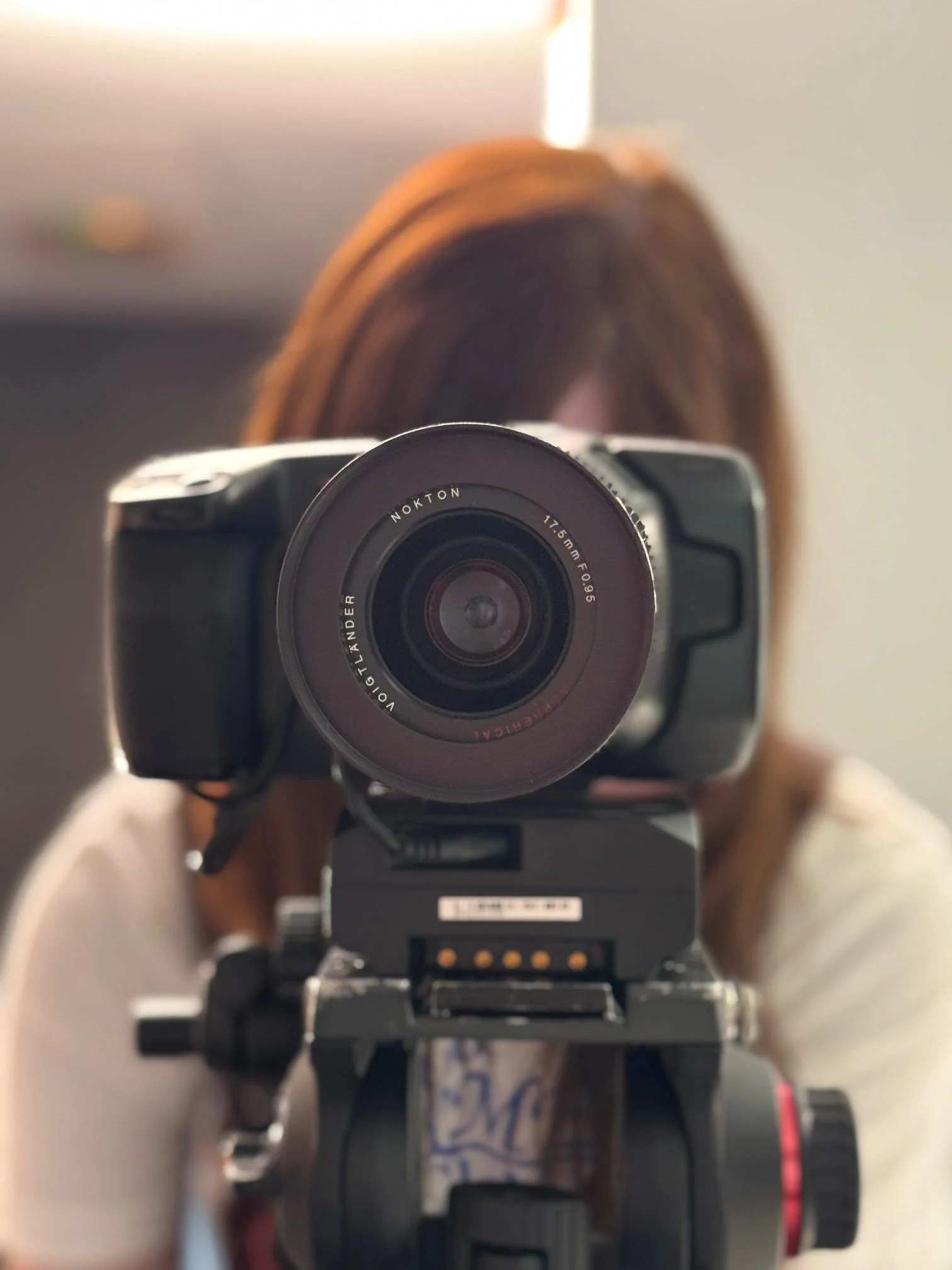 A person is taking a mirror selfie with a digital camera, capturing their face and upper body. The camera lens and person's hair are visible in the reflection.